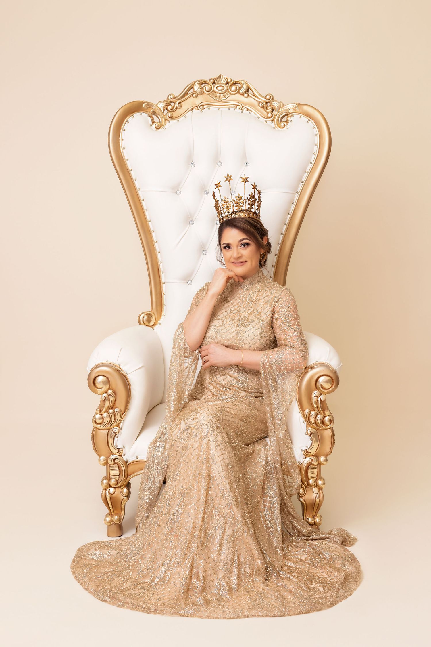 Elegant portrait in beige lace gown and crown seated on ornate white and gold throne.