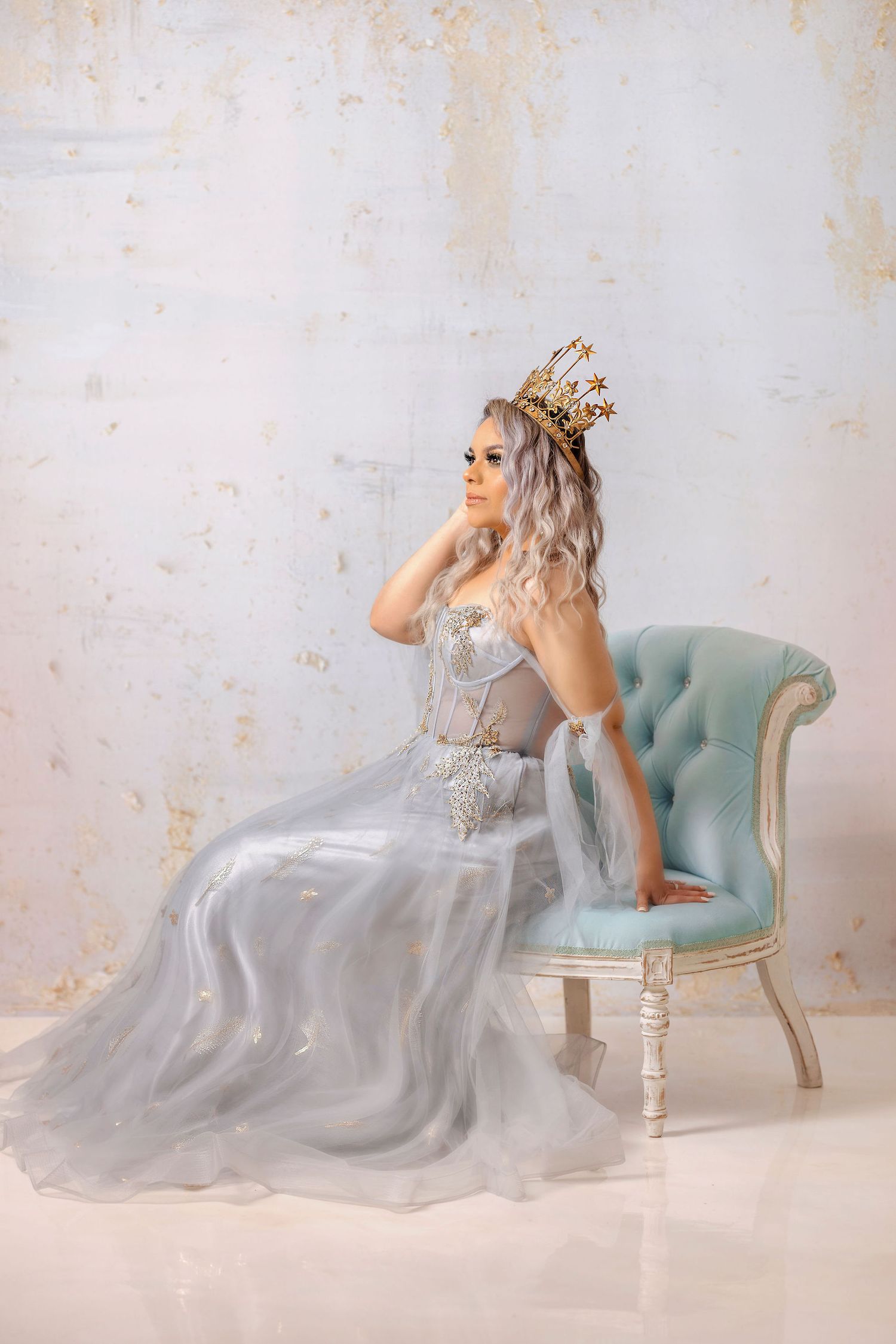 Ethereal silver ball gown and crown posed on vintage blue chair against textured wall.