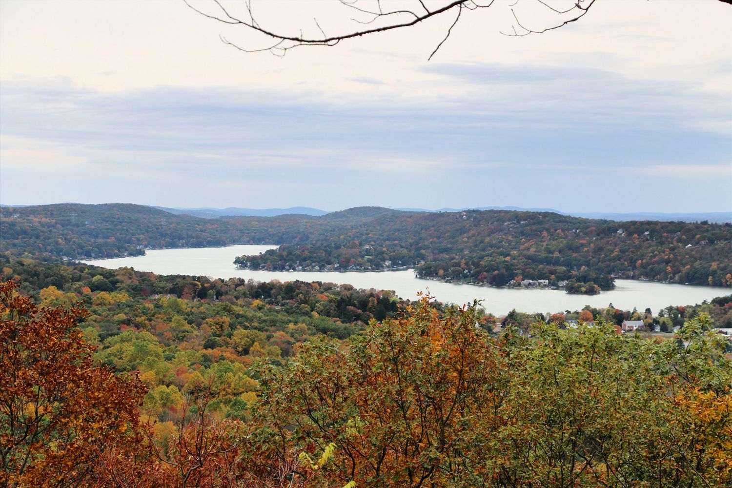 Aerial view of lake surrounded by vibrant fall foliage and rolling hills under cloudy sky.