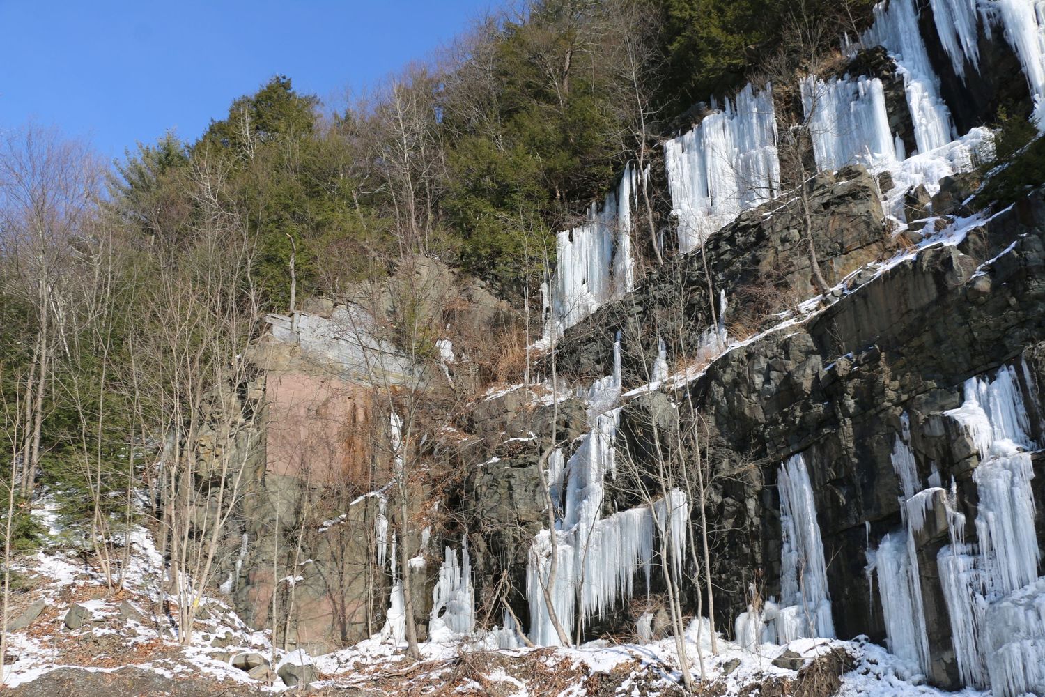Dramatic frozen ice formations cascading down rocky cliff face in winter forest landscape.