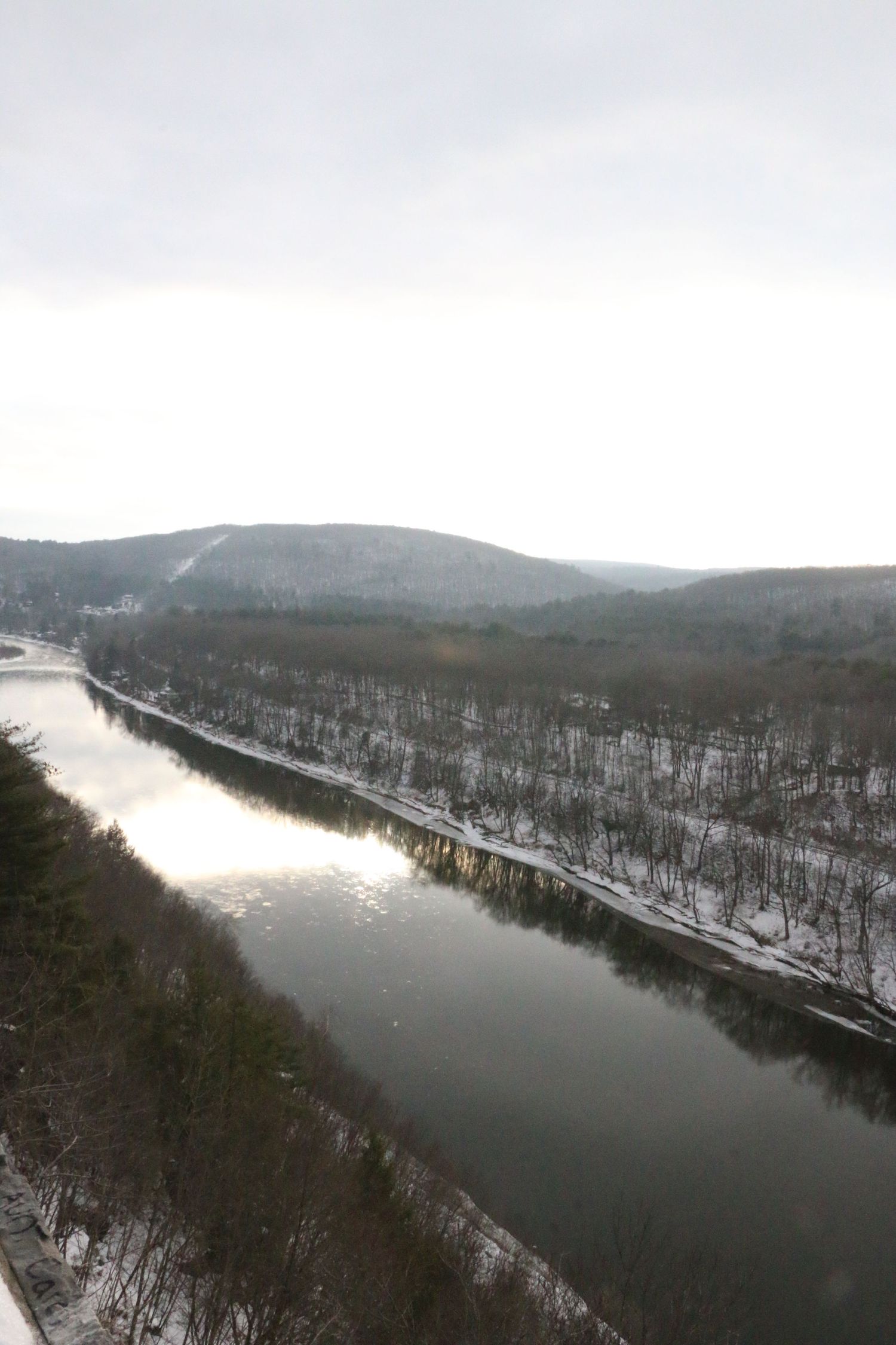 Aerial winter view of calm river reflecting light between snow-covered forested hillsides.