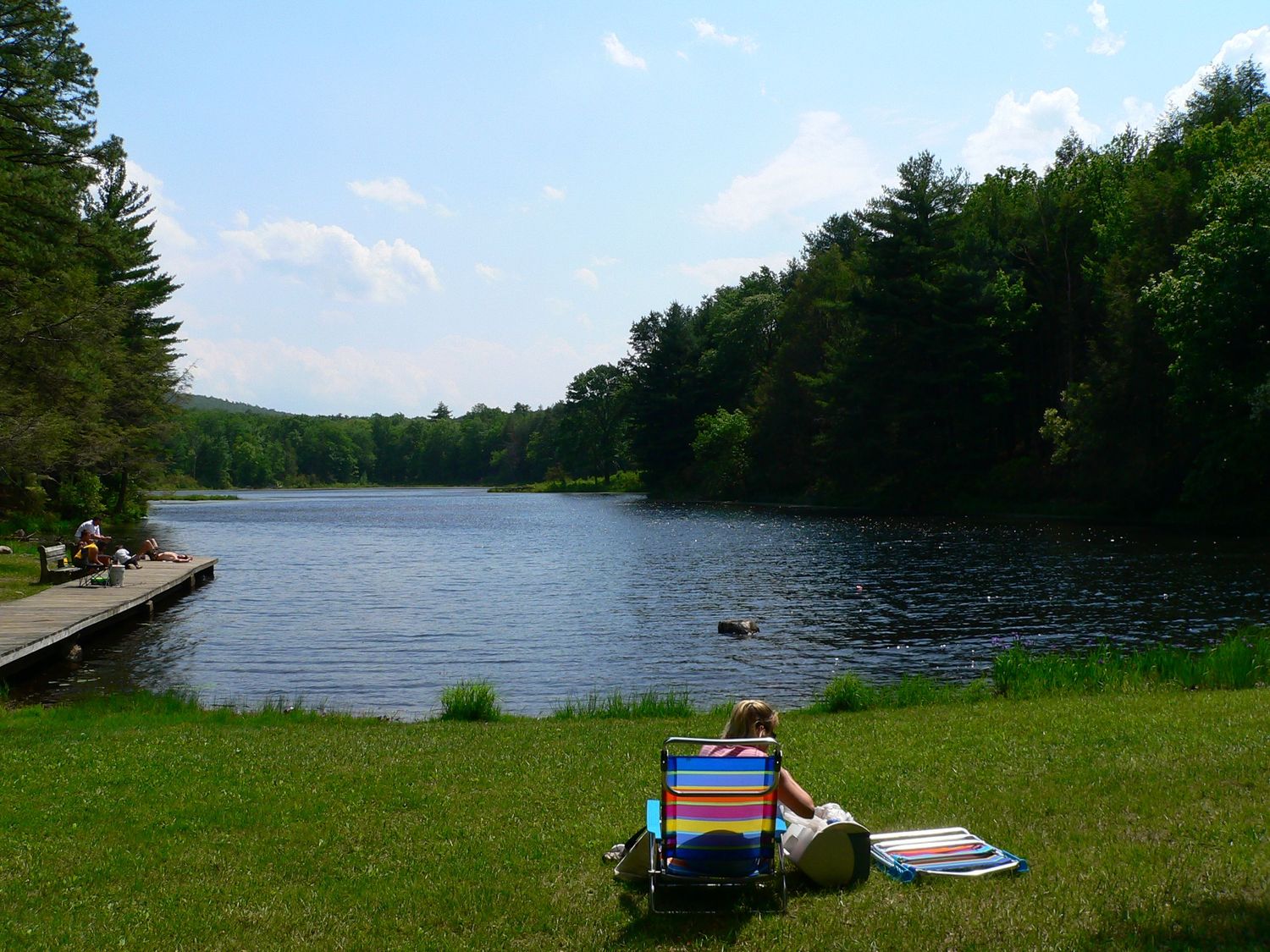 Person relaxing in lawn chair by peaceful forest lake with wooden dock on sunny day.