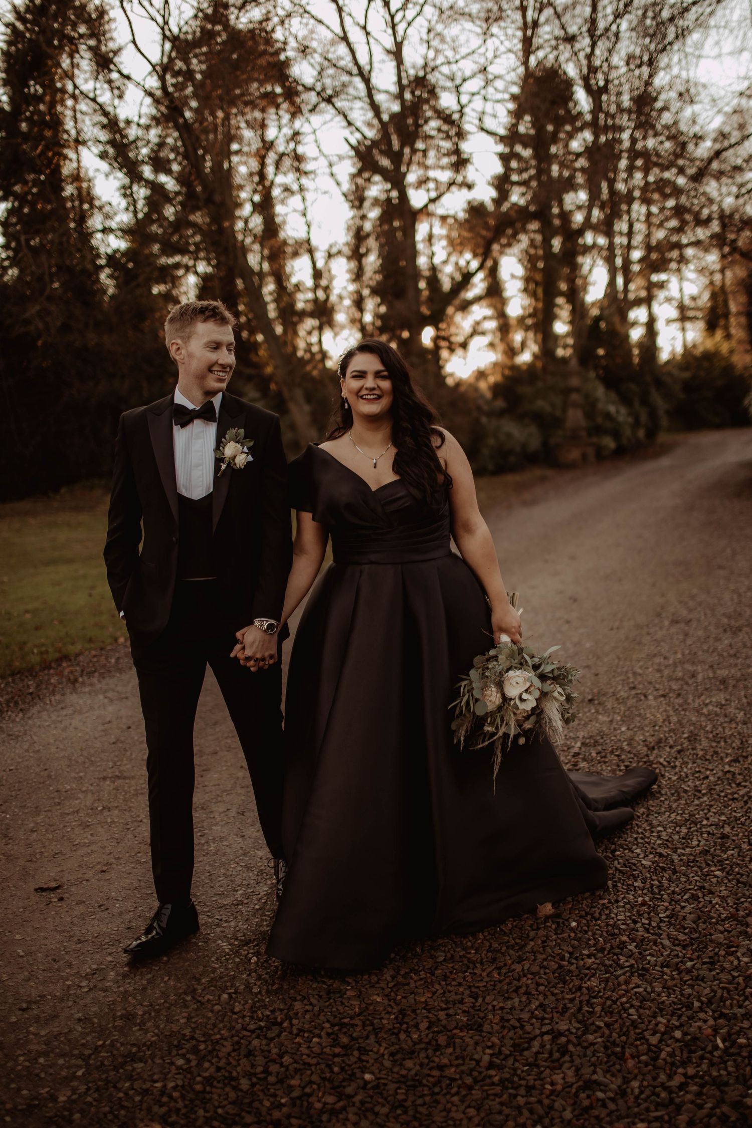 Couple walking down autumn path during sunset, bride wearing elegant black gown.