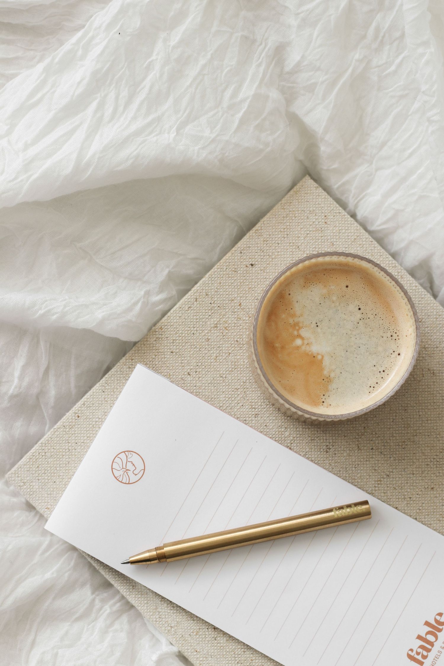 Minimalist workspace with coffee cup, gold pen and blank notepad on textured white background.