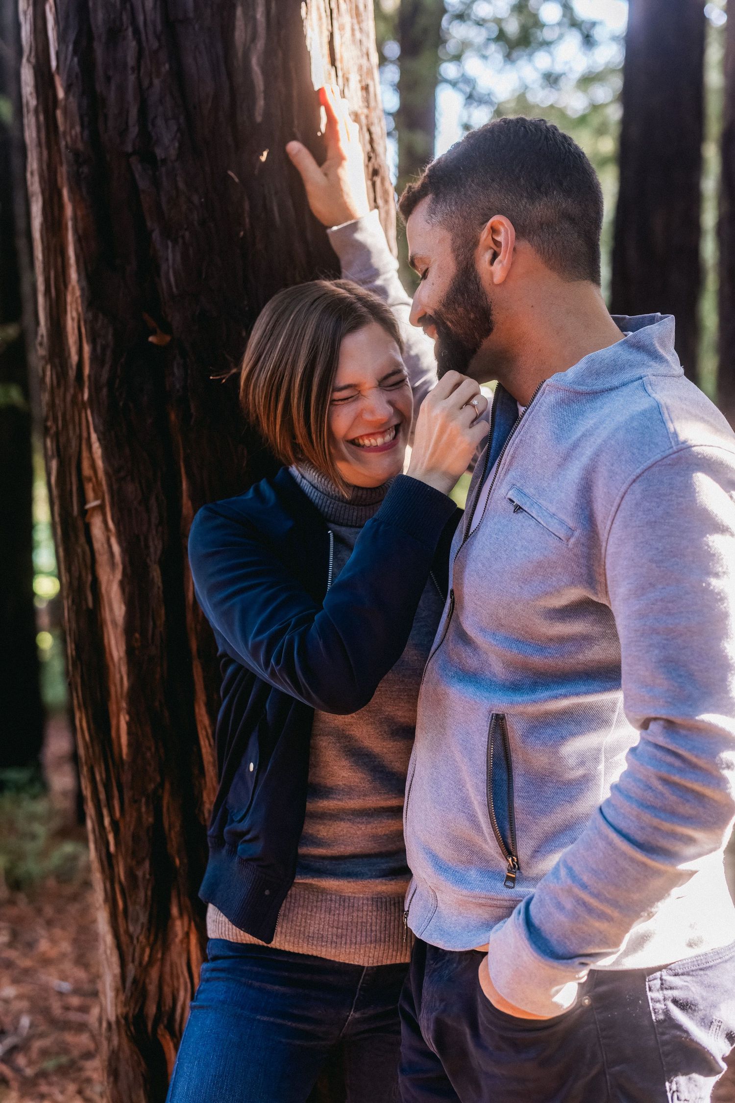A joyful couple shares a laugh while leaning against a large redwood tree.