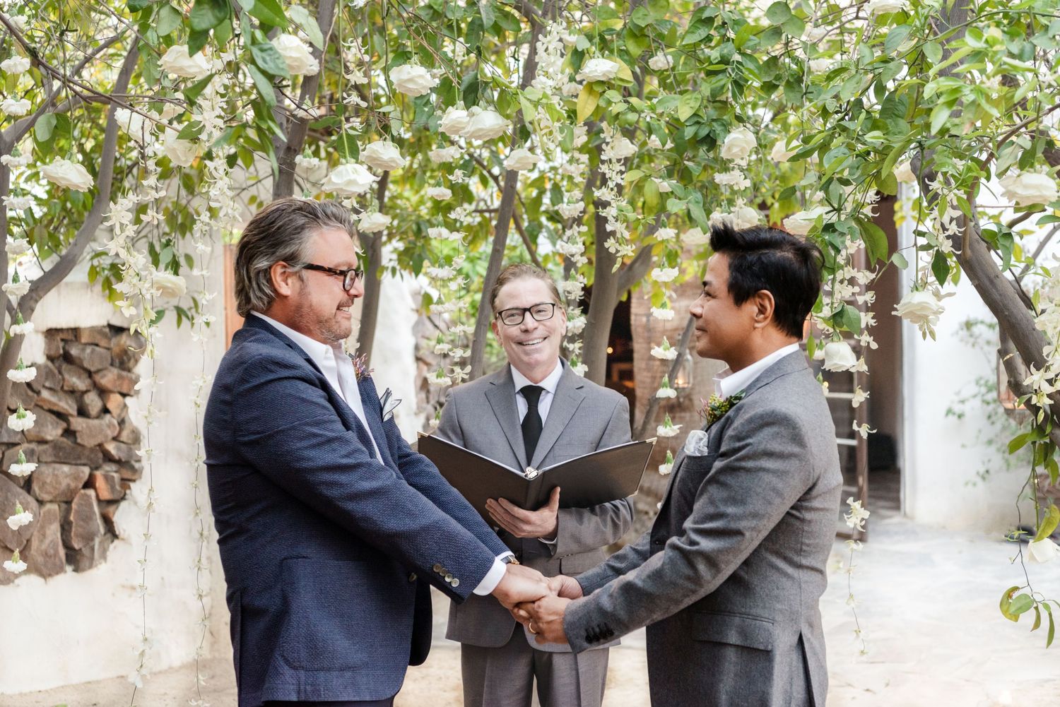 Three people in suits participating in outdoor ceremony under flowering trees.