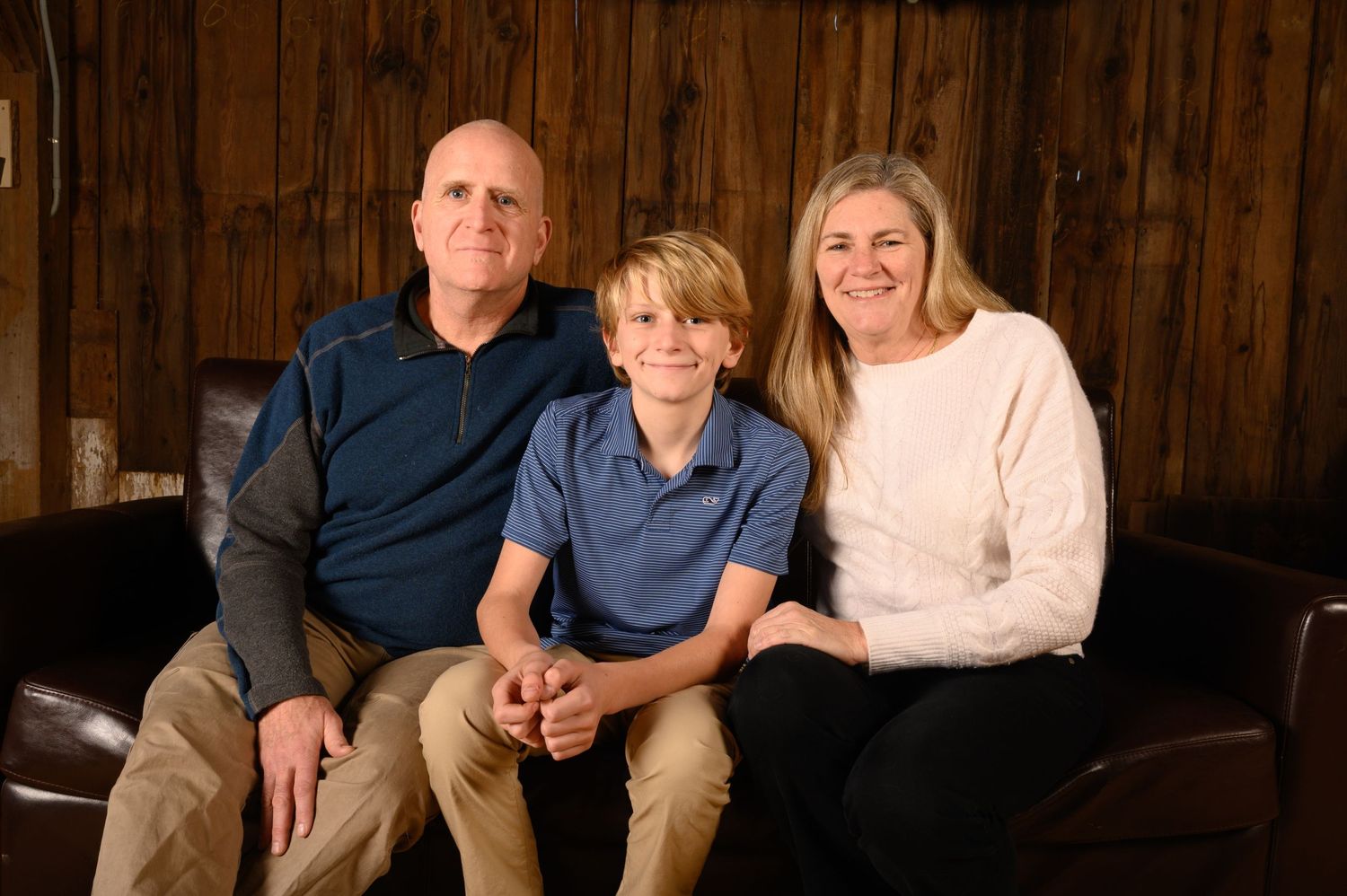 Casual family portrait with three people on leather couch in rustic wood-paneled room.