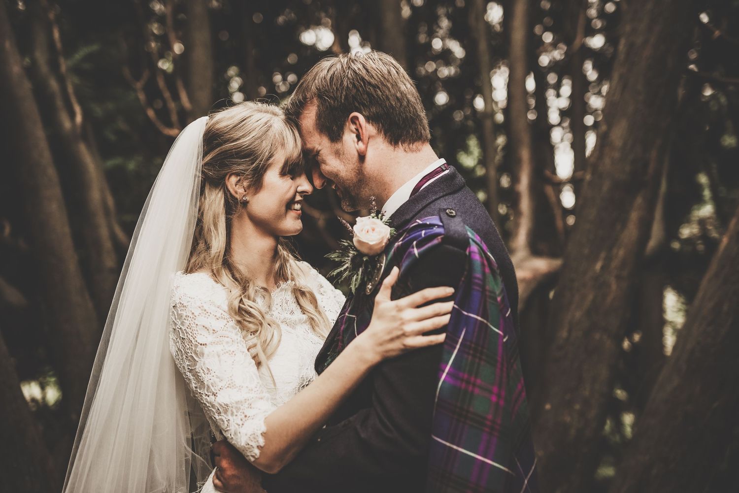Romantic wedding portrait of couple embracing in a wooded setting with bride in lace dress and groom in plaid suit.