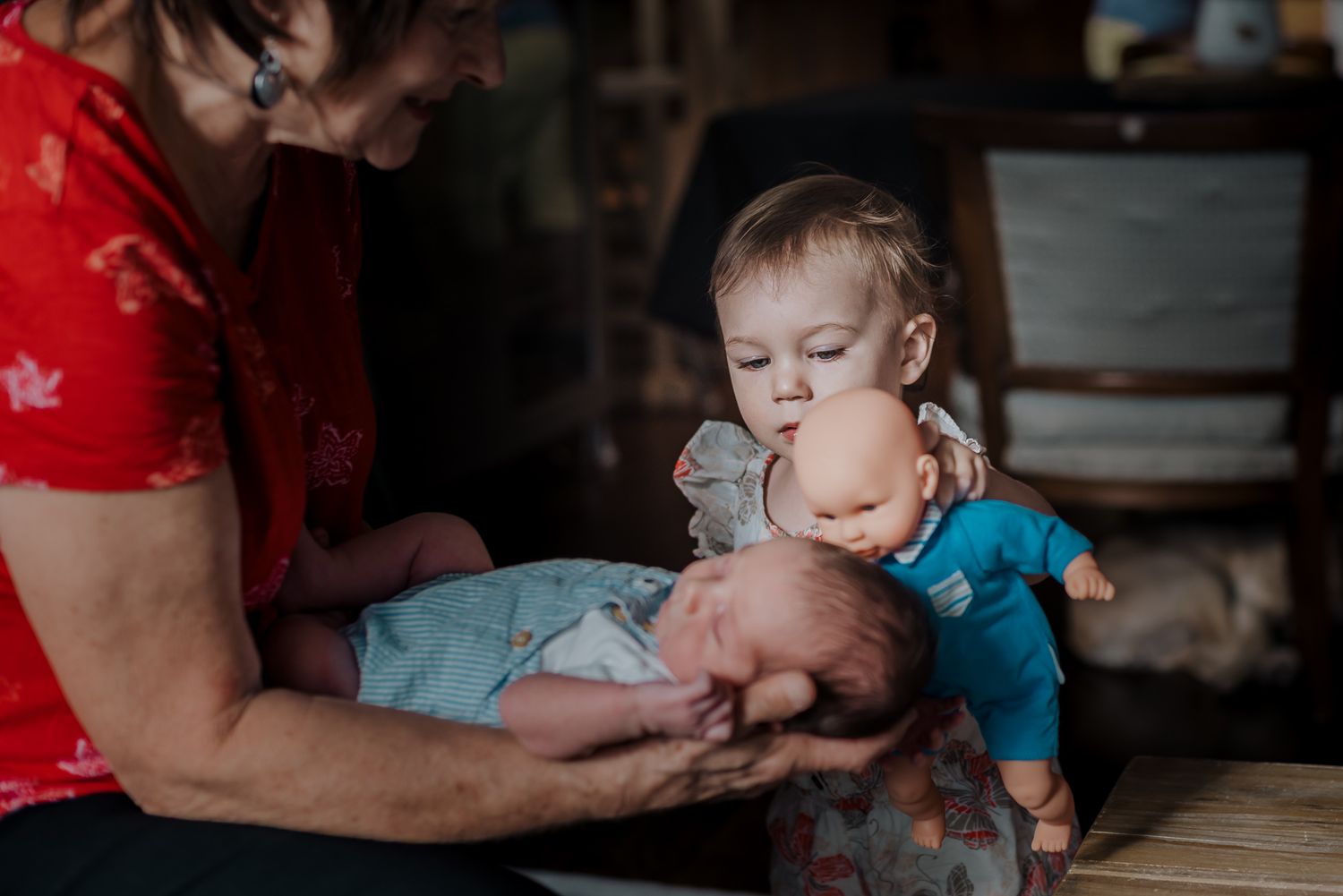 A toddler holds her baby doll up to her newborn sibling who's being held by his grandmother.
