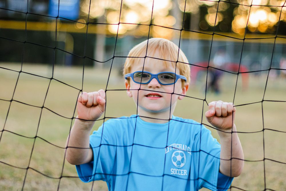 Young player in light blue jersey smiles while holding onto a soccer goal net.