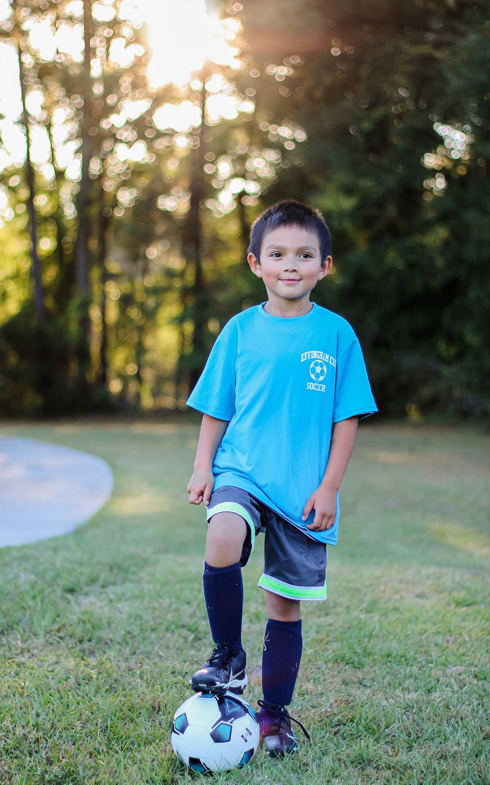 Young soccer player in light blue shirt and navy shorts poses with soccer ball at sunset.
