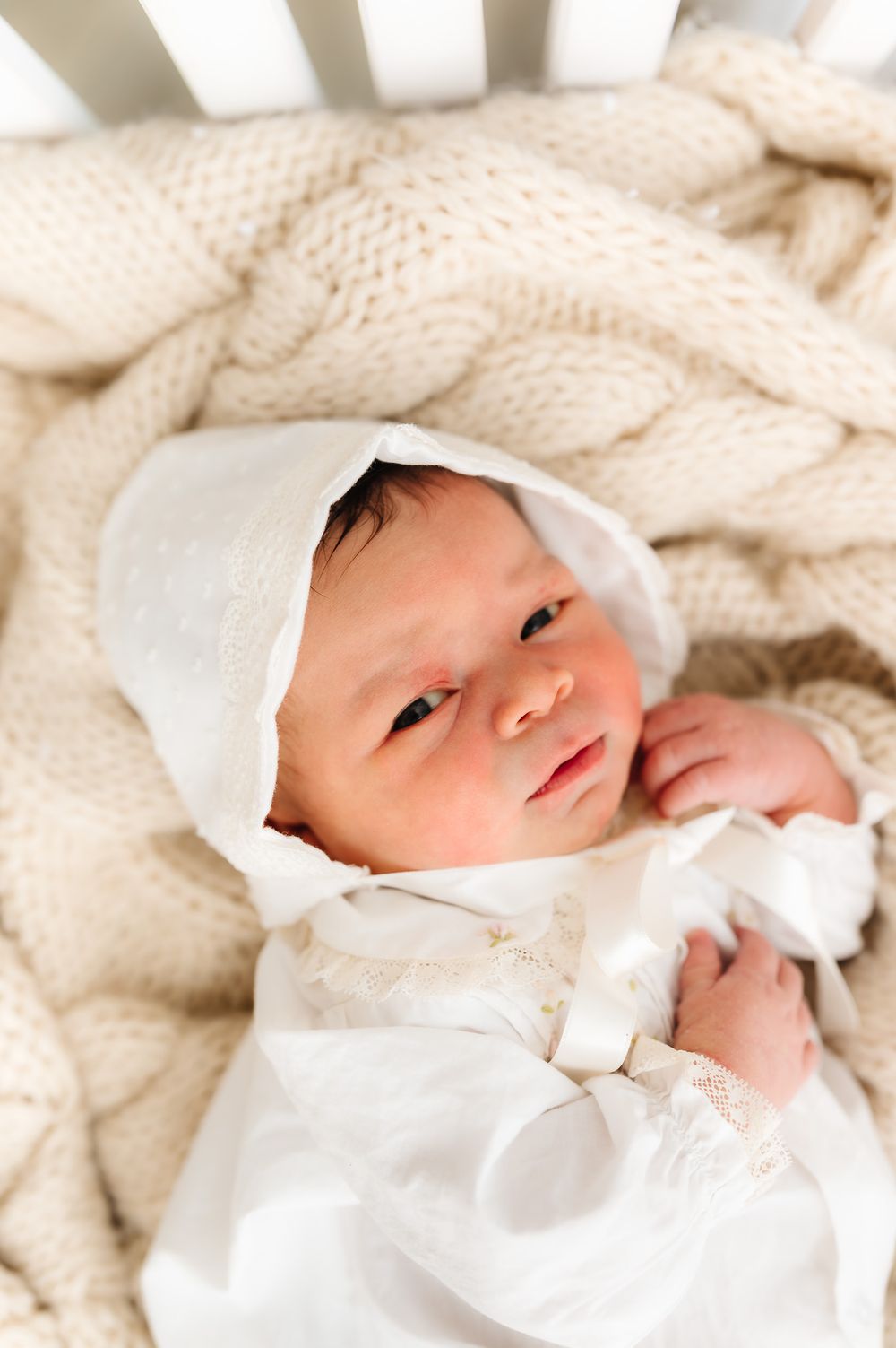 Newborn baby wearing white bonnet resting on cream knit blanket.