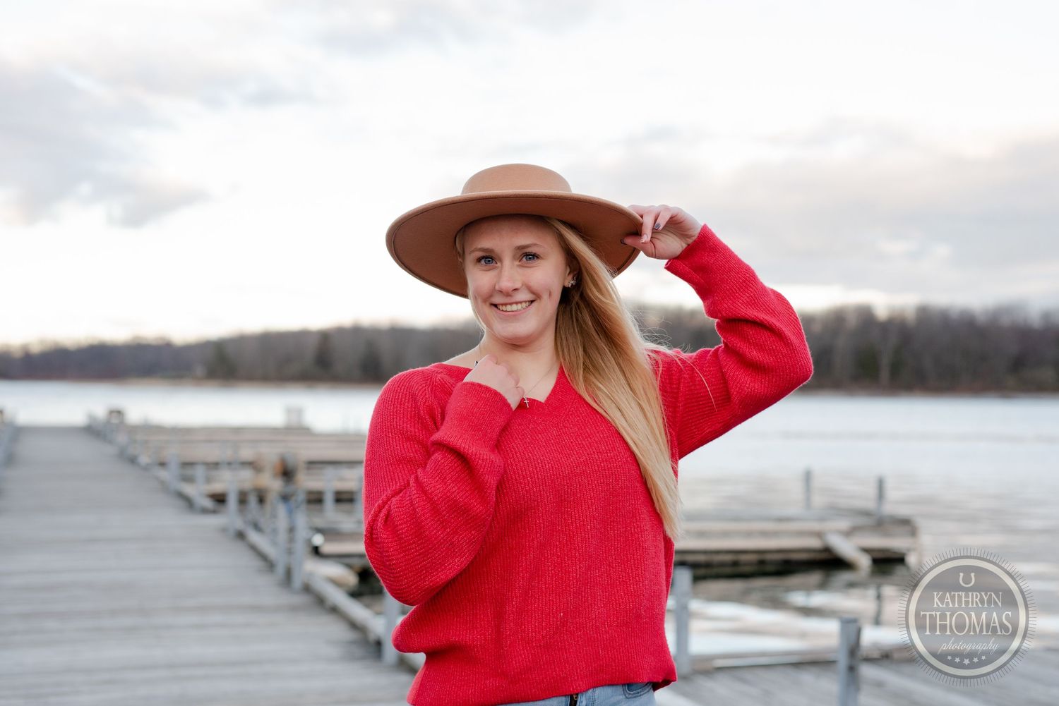Person in bright red sweater and brown hat posing cheerfully on waterfront dock.