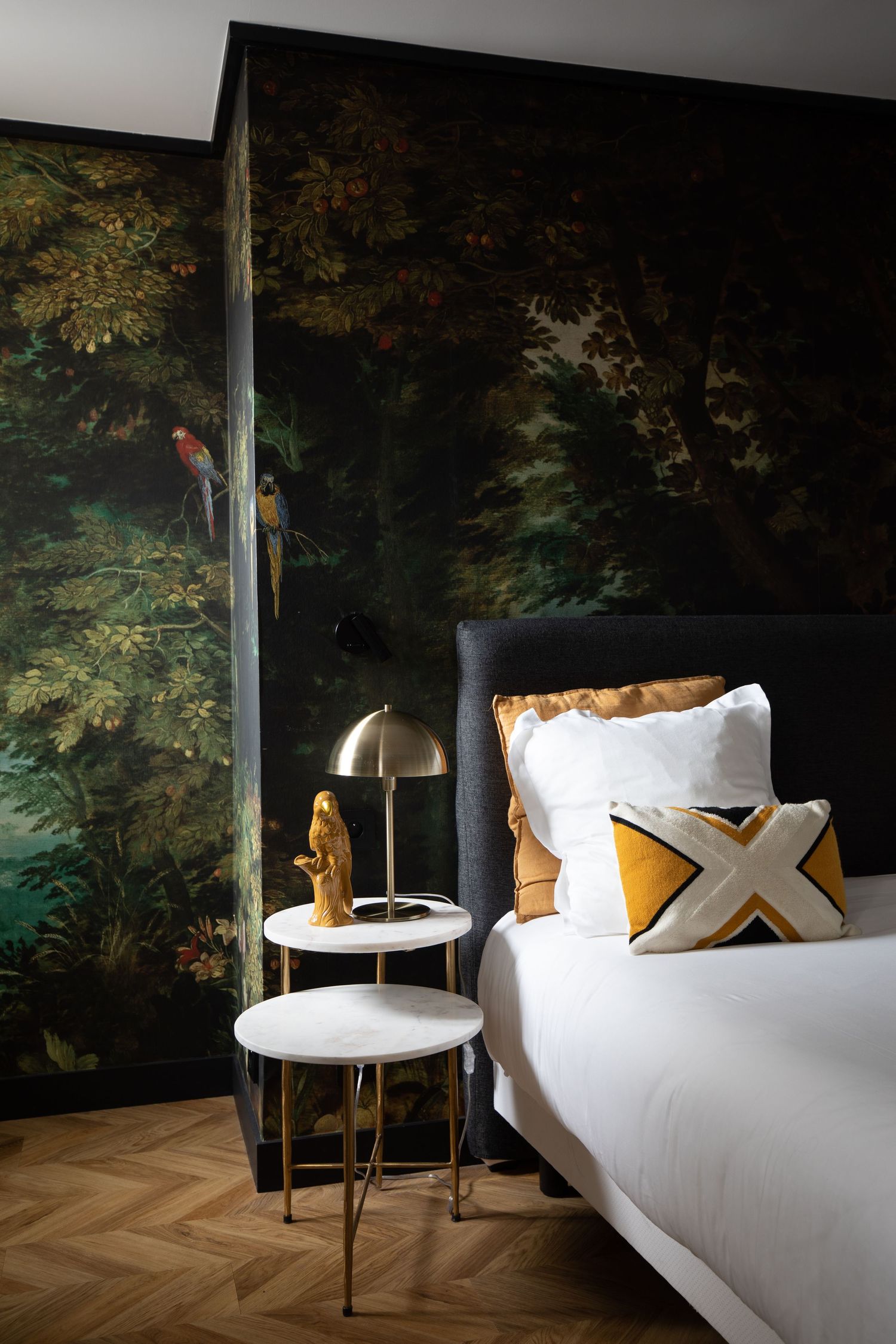 Elegant bedroom with forest wallpaper, herringbone floors, and modern side table with brass lamp.