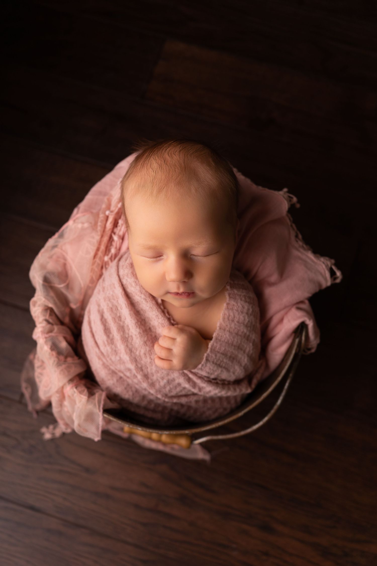 Newborn photography series showing peaceful sleeping baby wrapped in pink fabric on dark wooden surface.