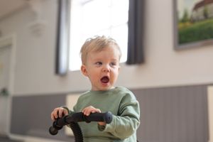 Excited toddler in green sweater grips a toy steering wheel in a playroom with grey walls and framed artwork.