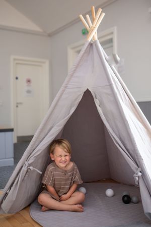 Child sitting cross-legged inside grey teepee tent with playful expression.