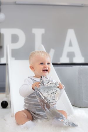 Joyful baby playing with silver foil in white and grey playroom setting.