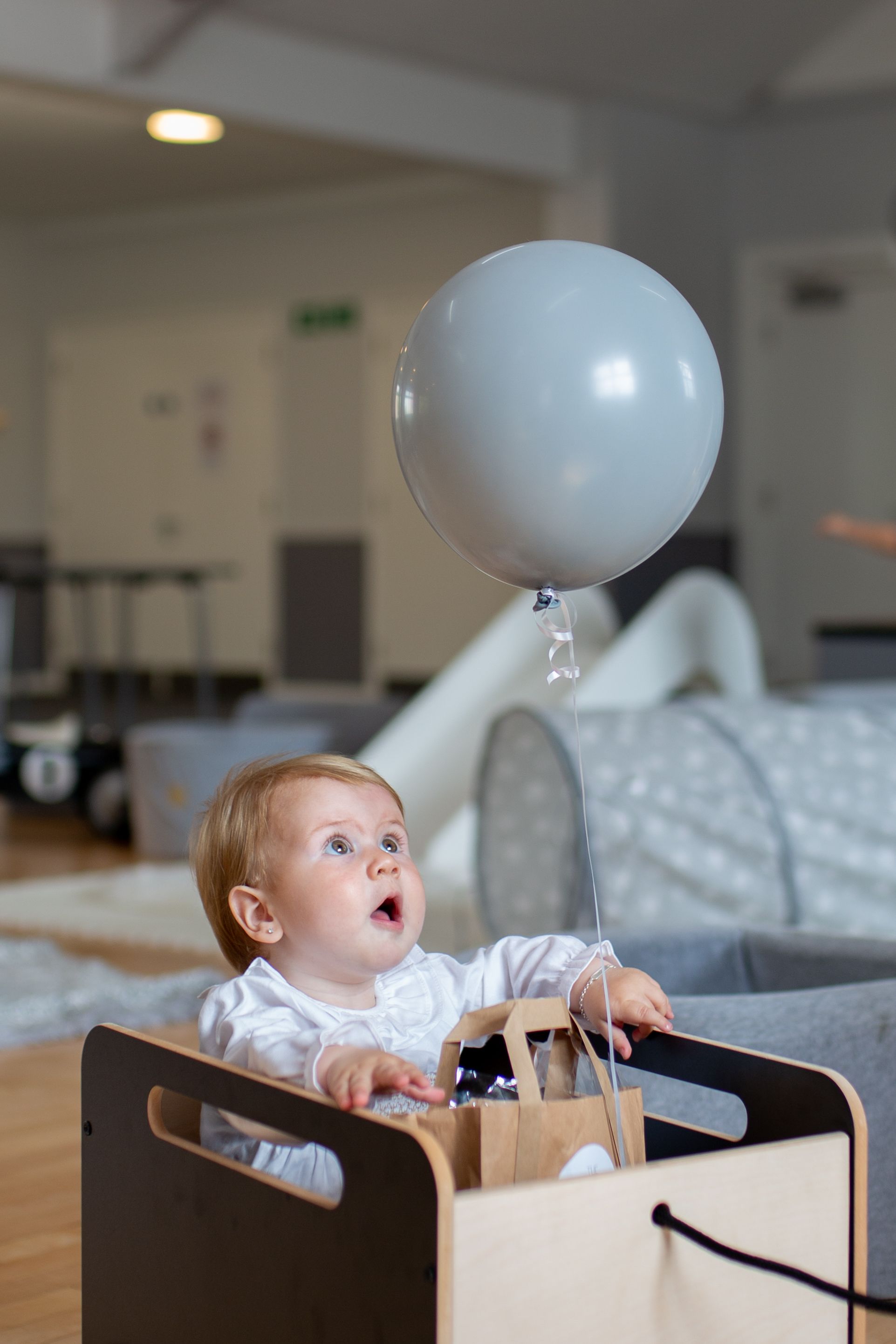 Baby in wooden toy car reaches for floating white balloon in modern playroom.