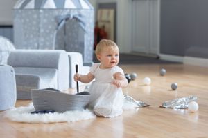 Small child in white dress plays with scattered toys on hardwood floor with grey decor.