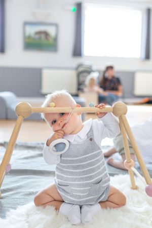 Baby in striped outfit playing with wooden activity bar on white fluffy rug.