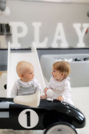 Two small children sit in black vintage-style ride-on car beneath PLAY lettering.