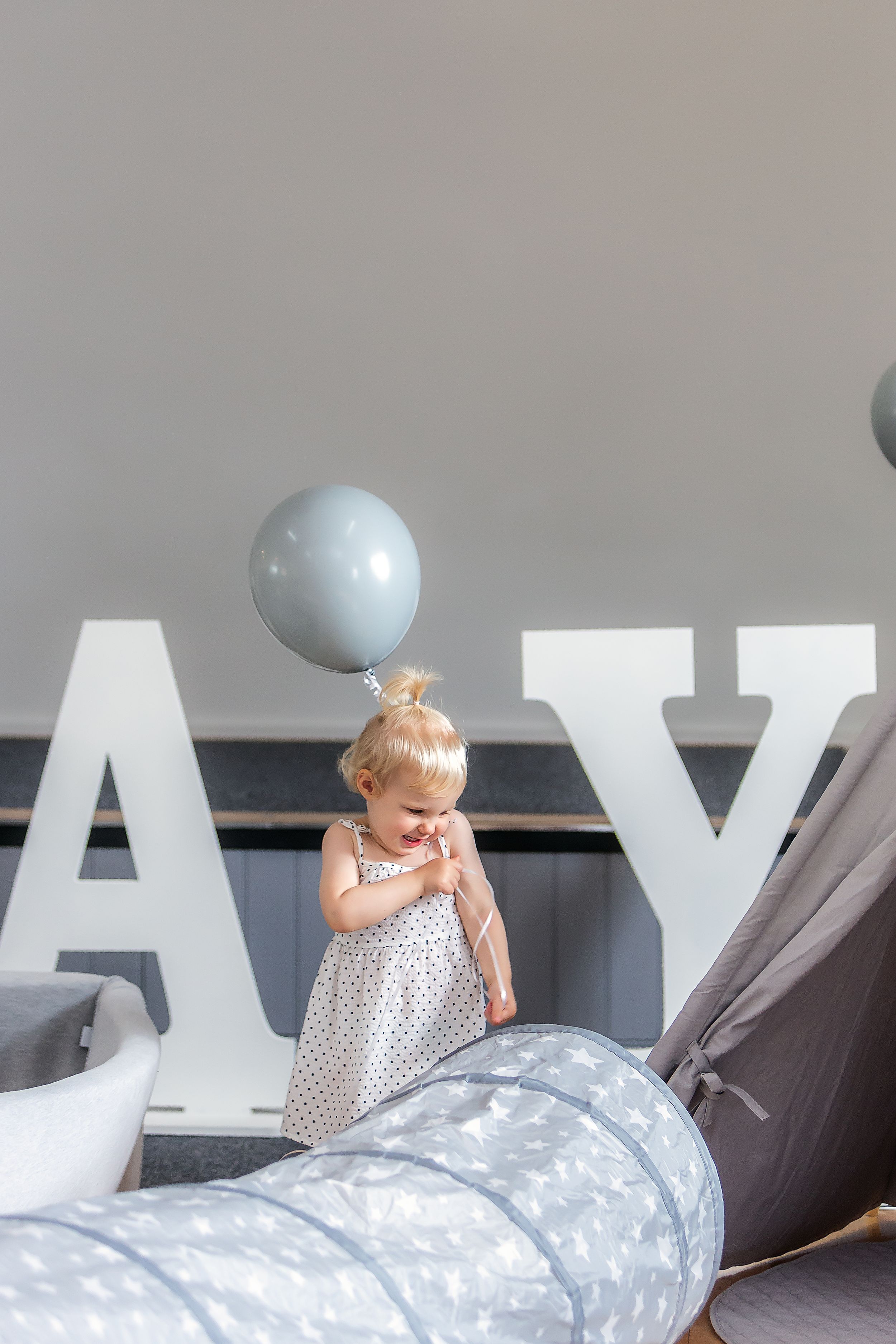 Toddler in polka dot dress standing by large decorative letters with balloon.