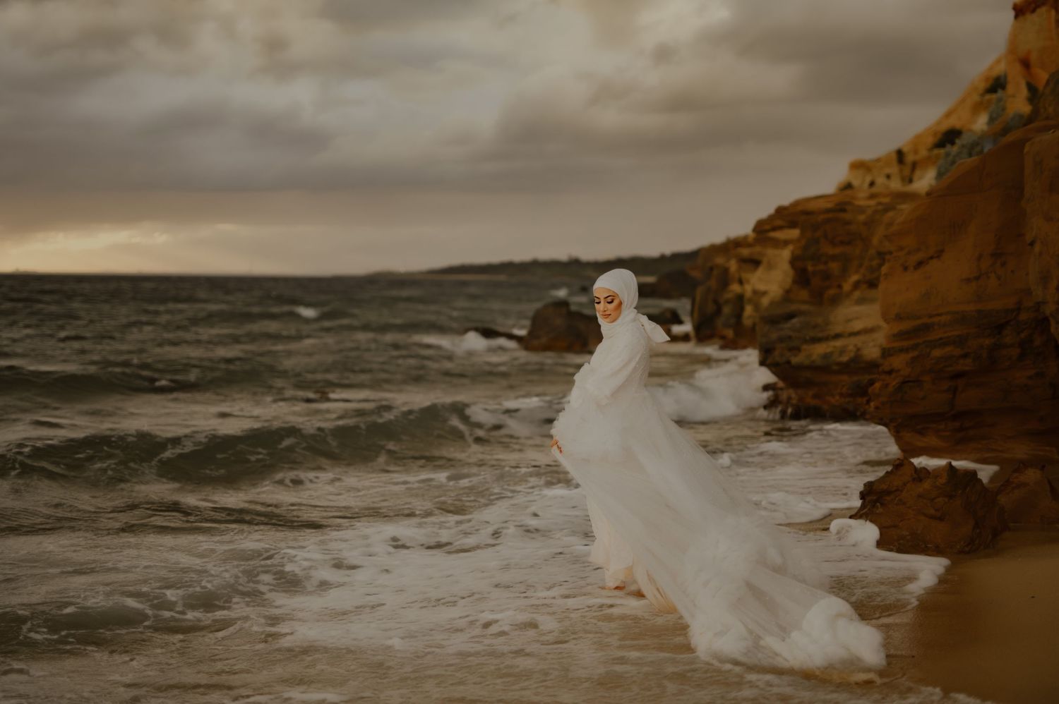 Dramatic coastal maternity photo with flowing white dress against stormy ocean backdrop.