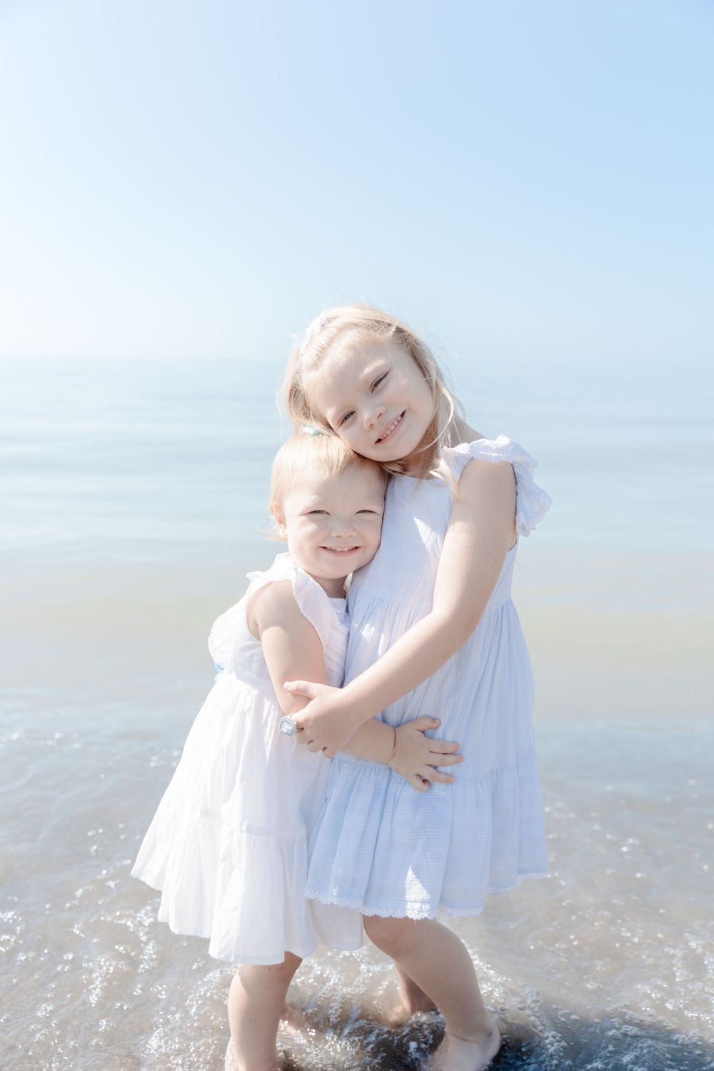 Two little girls in white dresses standing in the ocean.