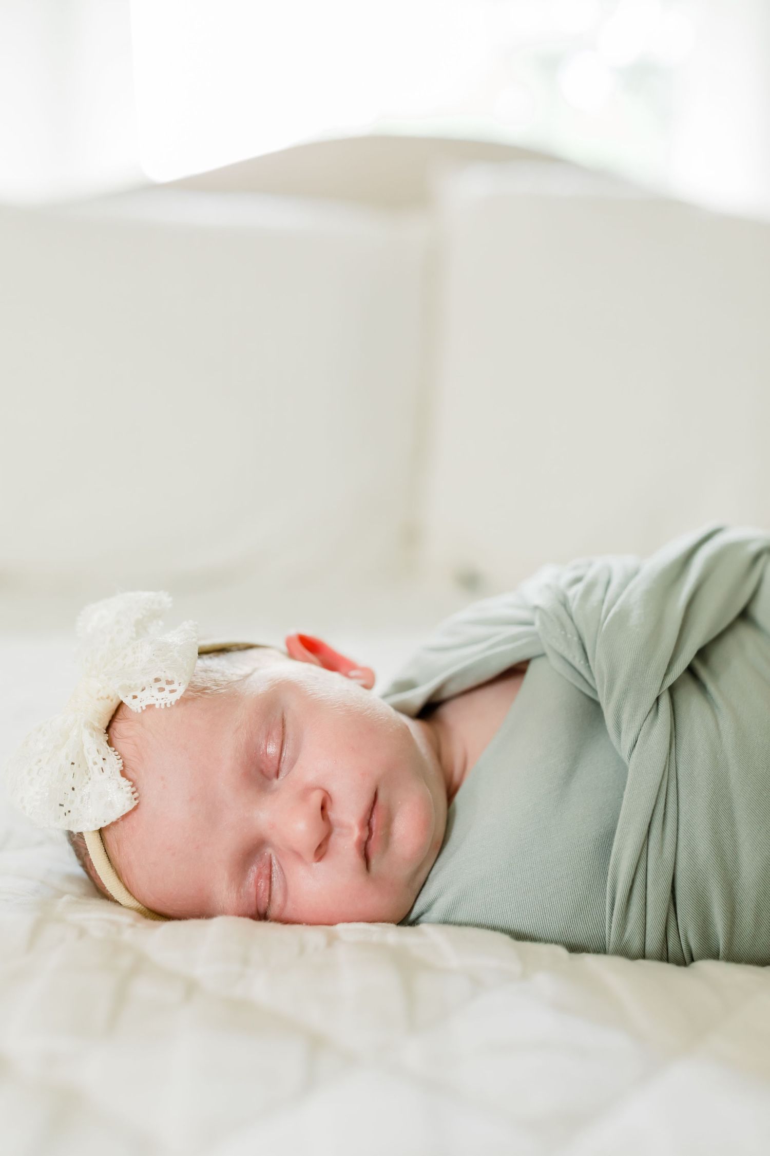 A sleeping newborn wearing a lace headband is wrapped in sage green fabric.