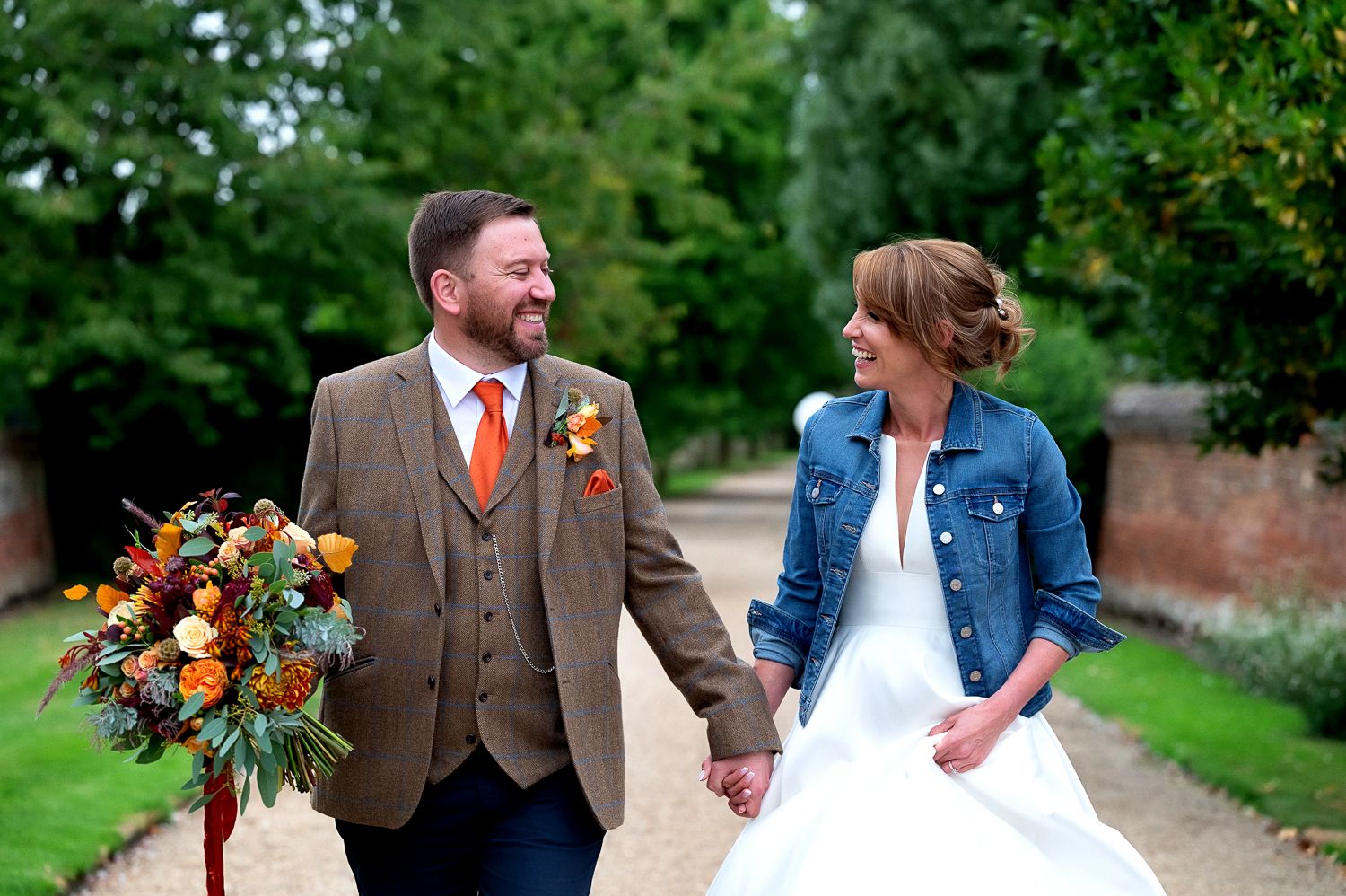 A couple wearing wedding attire walks together on a garden path while holding a vibrant autumn bouquet.