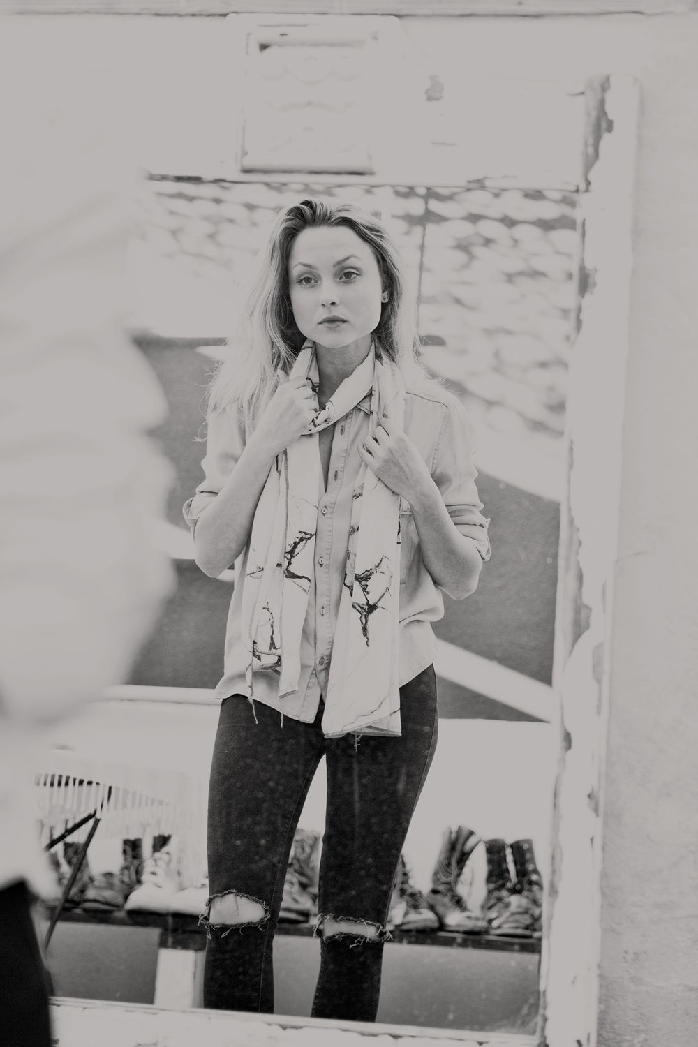 Black and white mirror reflection of a blonde woman wearing a printed scarf and ripped jeans.