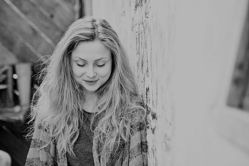 Young blonde woman leaning against weathered wall looking down in black and white portrait.