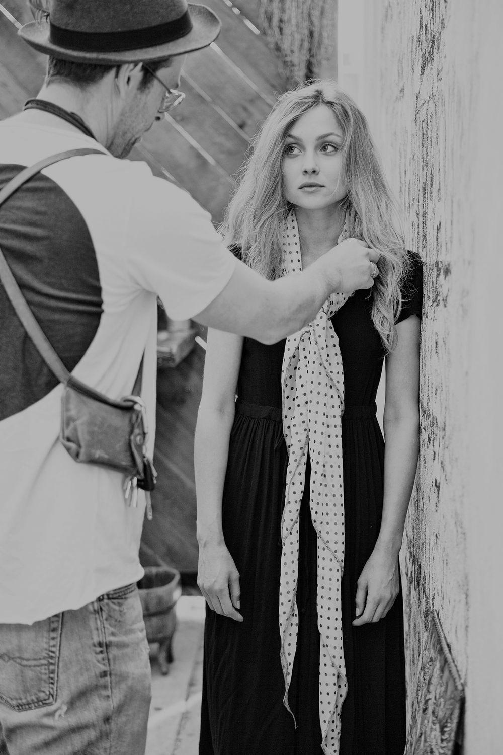Black and white photo of a man adjusting a polka dot scarf on a blonde woman outdoors.