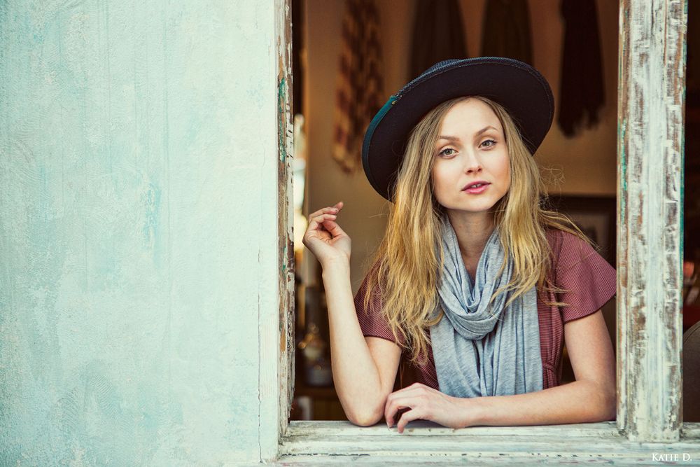 Blonde woman in wide-brim hat and gray scarf leaning out a rustic window.