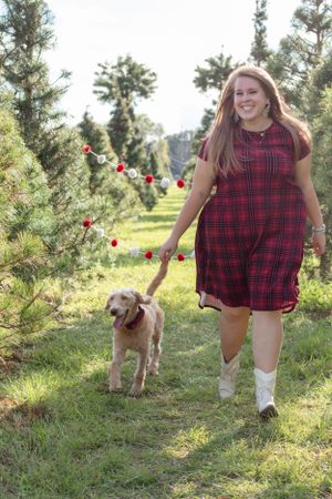 A woman in a red plaid dress walks with her golden retriever through a Christmas tree farm.