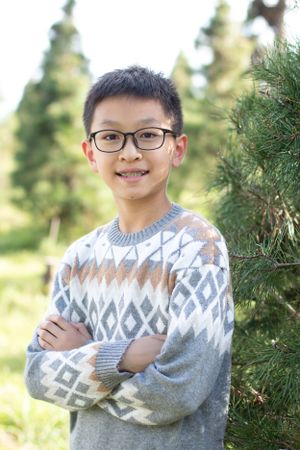 Young person wearing geometric patterned sweater poses near evergreen trees.