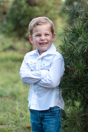 A child in white shirt and jeans poses confidently against an evergreen tree outdoors.