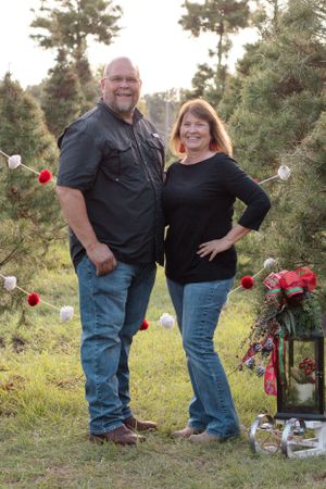 Couple posing by decorated Christmas trees with red ornaments and lantern in outdoor setting.