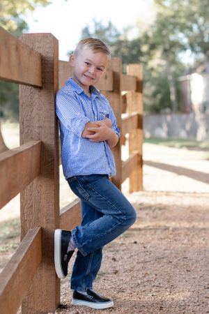 Young boy in blue checkered shirt poses casually against wooden fence outdoors.