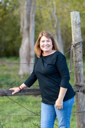 Woman in black sweater and jeans leaning against rustic fence post in outdoor country setting.