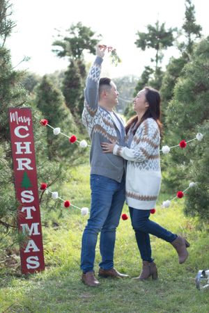 Couple in coordinating sweaters shares romantic moment by Christmas sign with red ornaments.