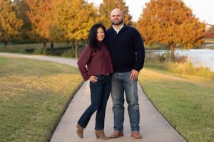 Couple poses together on a walking path with autumn trees and a lake in the background.