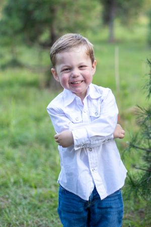 A candid portrait of a young child in white shirt and jeans laughing next to an evergreen tree.