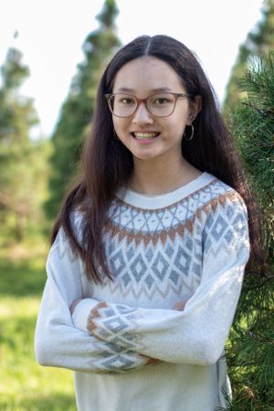 Person in fair isle sweater poses with crossed arms near evergreen trees.