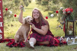 A woman in plaid dress sits on a blanket with her golden retriever among holiday decorations.