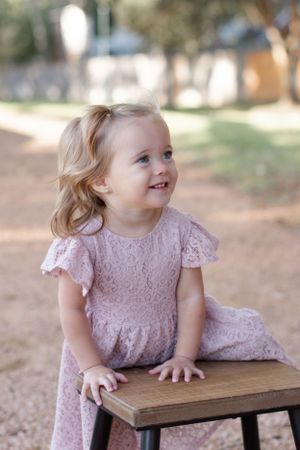 Young toddler in pink lace dress smiling while sitting on wooden stool outdoors.
