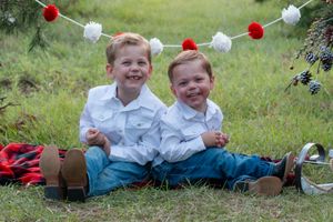 Two small children sit on grass under red and white holiday garland wearing matching white shirts and jeans.