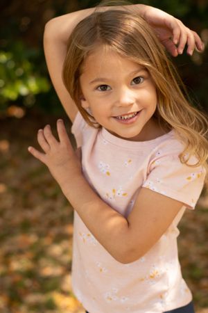 A playful child poses with hands raised wearing a light pink t-shirt against a natural backdrop.