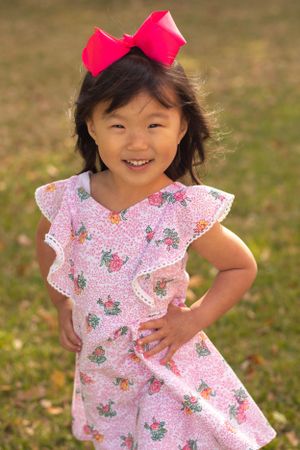 A child in a pink floral dress and red bow strikes a confident pose outdoors.